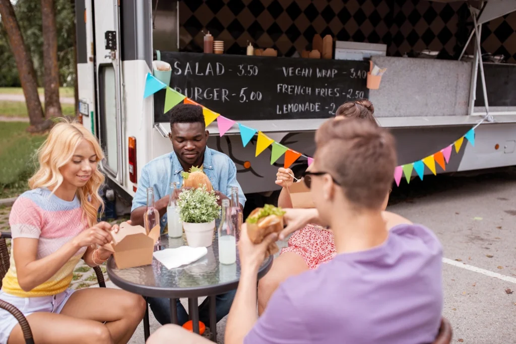 Group of friends enjoying burgers and compostable takeaway food at an outdoor table beside a colourful food truck during summer.