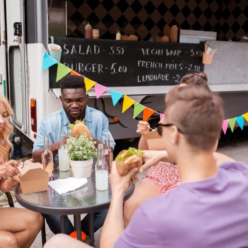 Group of friends enjoying burgers and compostable takeaway food at an outdoor table beside a colourful food truck during summer.